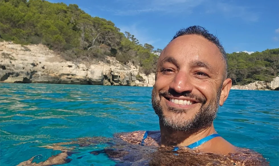 Fadi Salah smiling in clear turquoise water off Menorca, with limestone cliffs and pine trees behind him.