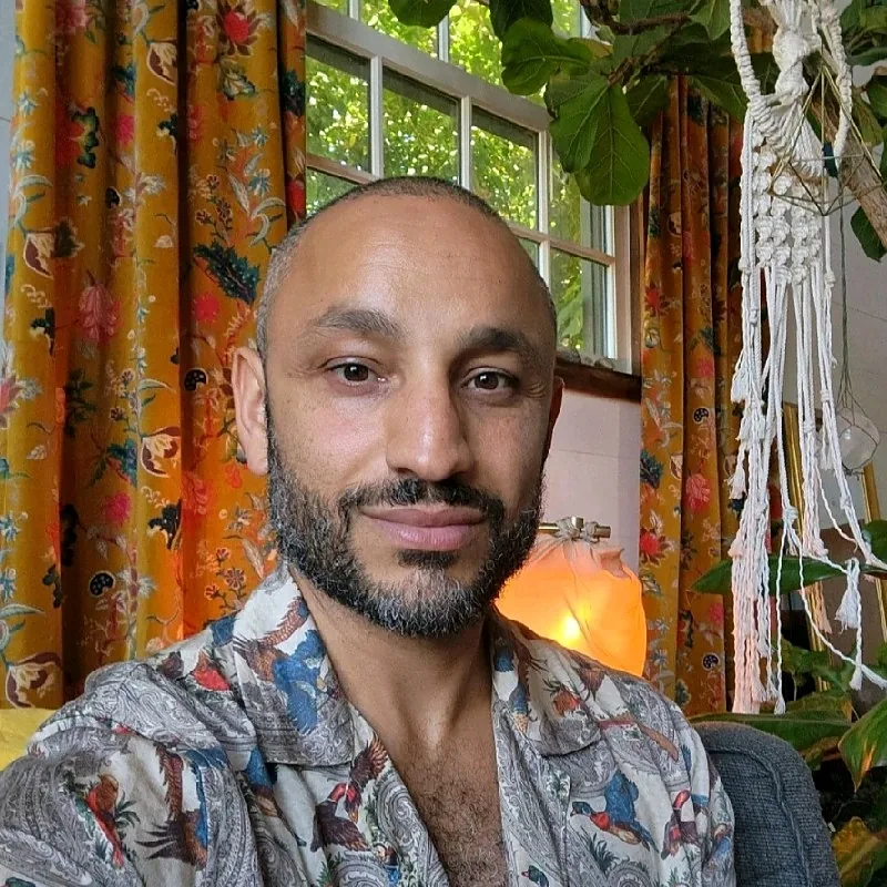 Fadi Salah, founder of Re:deem Atelier, at home against ochre floral curtains and warm lamplight.